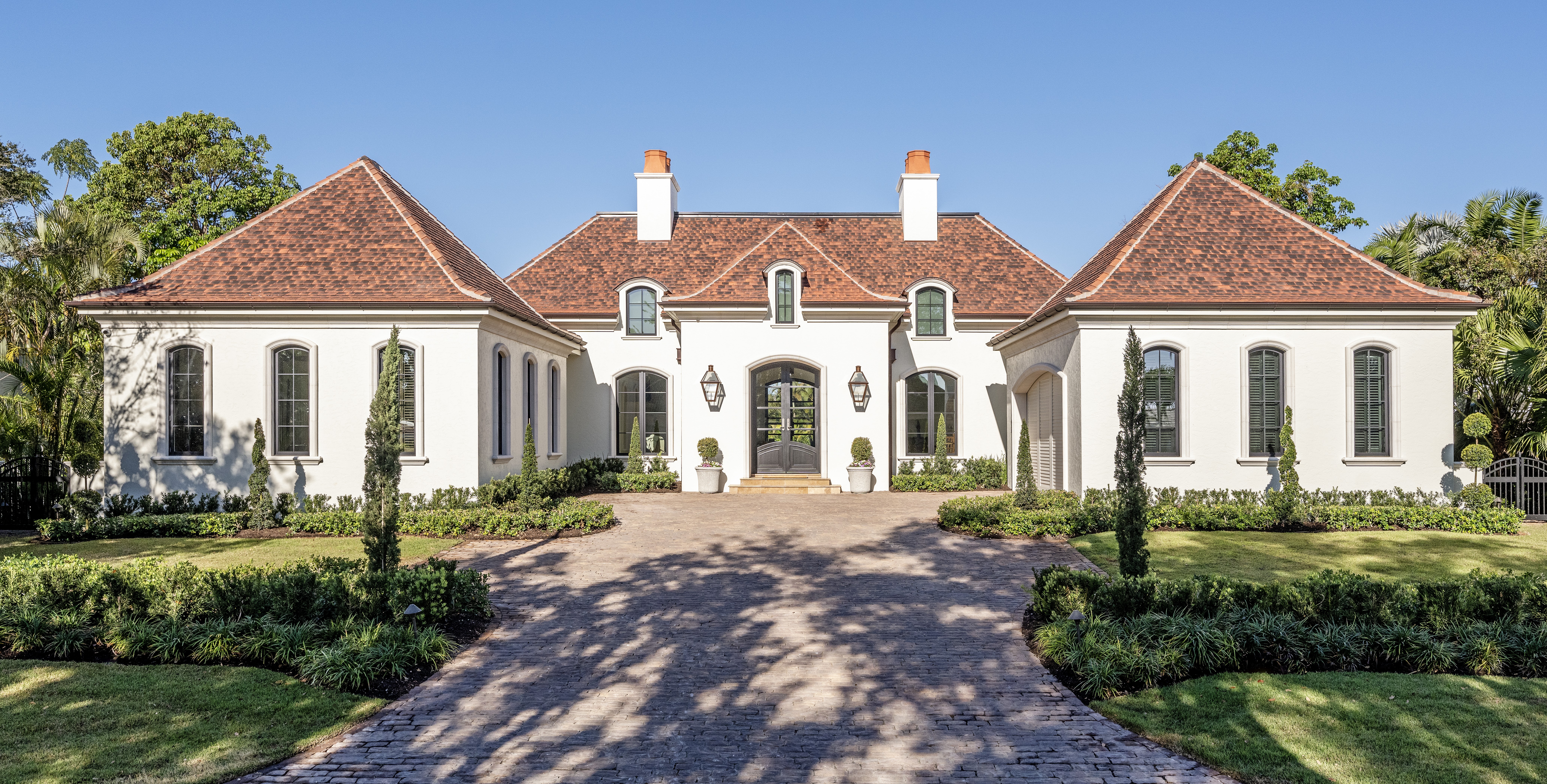 French Country-style residence in Sarasota by CMSA Architects, with symmetrical stucco façade, arched dormer windows, steep hipped red roofs, and a formal brick entry drive